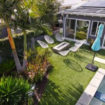 A photo of a beautiful backyard area with six chairs, an umbrella, artificial turf grass, afire pit, and a pool.